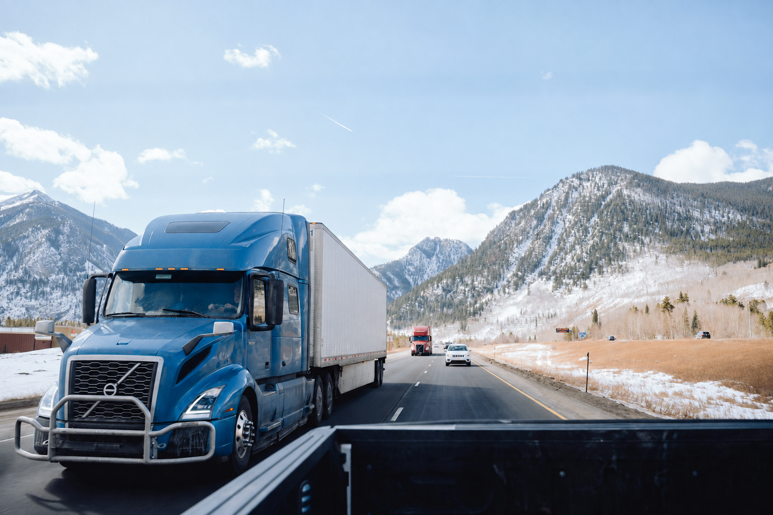 Blue American Volvo semi-truck with an enclosed trailer on a snowy mountain highway